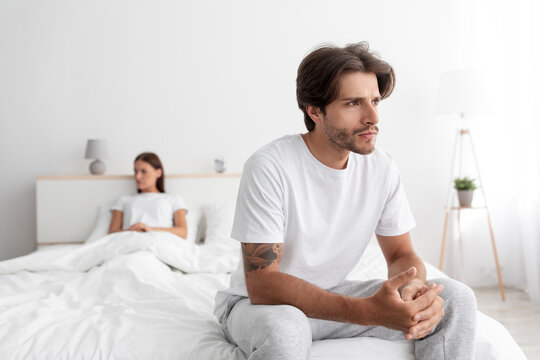 Upset sad offended millennial european husband and wife sitting on bed ignoring each other in white bedroom interior