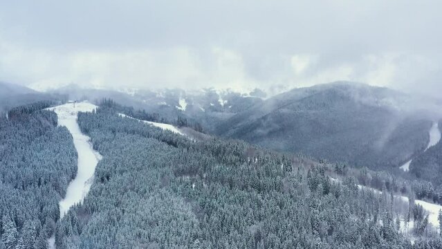 Aerial beautiful winter mountain landscape. Drone moving above the snow capped pine forest in moody weather above ski resort