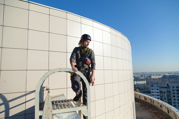 A worker in a helmet and climbing gear stands on the roof of a building and looks into the distance. Industrial climber during work