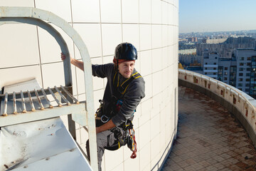 A self-confident industrial climber in gear holds onto a ladder on the roof of an office building