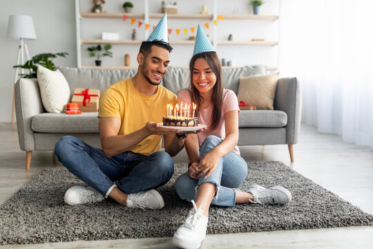 Young international couple in festive hats sitting on floor with birthday cake, celebrating together in living room - Powered by Adobe