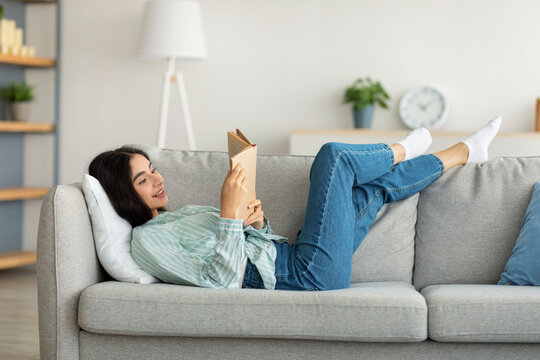 Stay home pastimes. Millennial Indian woman lying on sofa with open book indoors, enjoying peaceful weekend morning