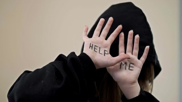 Teenage girl, covering her face with a hood, shows her palms with the inscription HELP ME.
