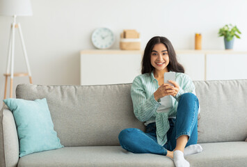 Fototapeta premium Charming young Indian woman sitting on sofa with smartphone, checking social media, chatting online at home