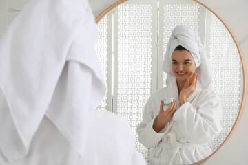 Beautiful young woman with hair wrapped in towel applying cream near mirror indoors