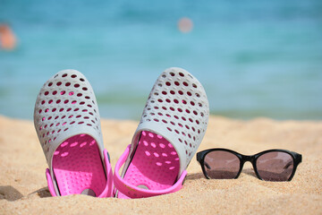 Closeup of clogs shoes and black protective sunglasses on sandy beach at tropical seaside on warm...