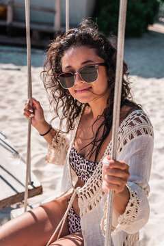 Vertical portrait of young hispanic woman with sunglasses and bikini sitting in a hammock on a caribbean beach