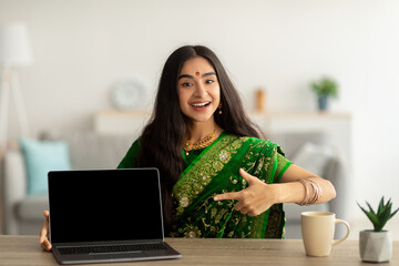 Cheerful Indian woman in green sari dress pointing at laptop computer with empty screen at home,...