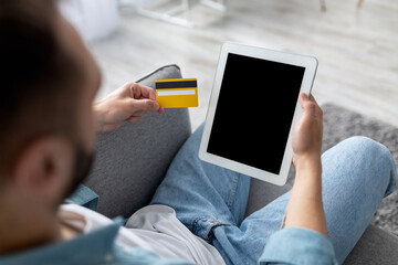 Unrecognizable young guy sitting on couch, holding credit card, shopping online on digital tablet at home, mockup