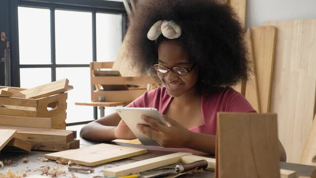 Medium shot portrait of a happy African-Thai teenager girl using digital tablet, smiling, laughing, lingering while studying carpentry class. Teen student and social media addiction concept.