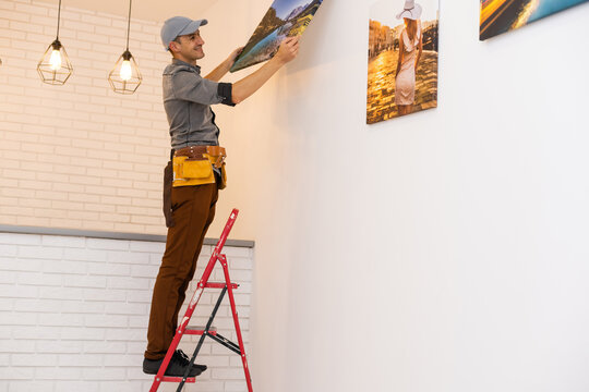 Young man standing on stepladder while his wife helping him to hang picture on wall in living-room of new flat or house