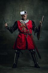 Portrait of man, medieval warrior, knight with dirty wounded face in VR headset isolated over dark vintage background. Comparison of eras, history