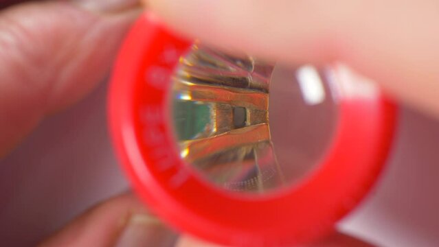 Closeup Overhead Shot Of Fingers Using A Magnifying Loupe To Closely Examine A Gold, Diamond And Emerald Ring.