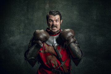 Fototapeta na wymiar Portrait of medieval warrior or knight with dirty wounded face in boxing gloves isolated over dark background. Comparison of eras, history