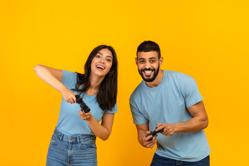 Having fun. Playful arab couple playing video games together, holding and using joysticks over yellow background © Prostock-studio