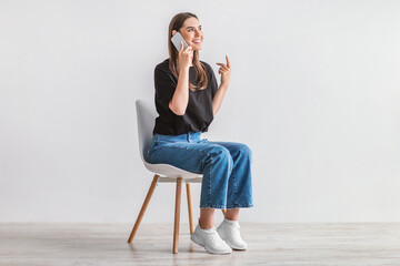 Smiling young woman having conversation on cellphone, sitting on cozy chair against white studio...