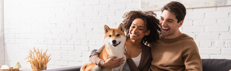 Happy man looking at smiling african american girlfriend with shiba inu at home, banner
