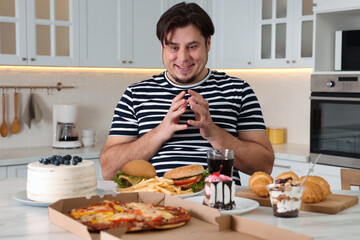 Fototapeta na wymiar Hungry overweight man at table with sweets and fast food in kitchen