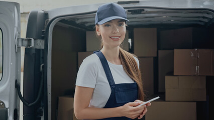 Cheerful delivery woman holding smartphone and looking at camera near blurred boxes in car outdoors