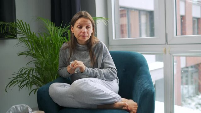 Unhealthy woman sits in a chair and sneezes or blows her nose into a napkin because she has a cold, flu, coronavirus. It is snowing outside