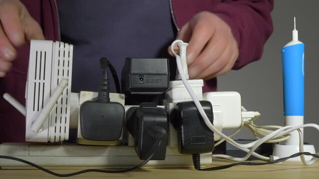 Closeup of a man’s hands holding an overloaded, UK 240 volt electricity supply extension board, with a mess of adaptors, wires and cables, while plugging in a wi-fi extender device.
