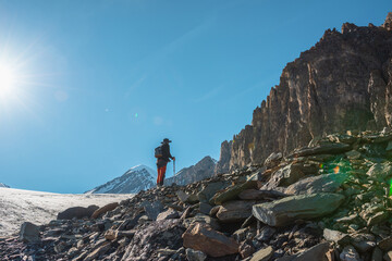 Fototapeta premium Scenic alpine landscape with silhouette of hiker with trekking poles against large sharp rocks and snow mountain peak in sunlight. Man with backpack in high mountains under blue sky in sunny day.