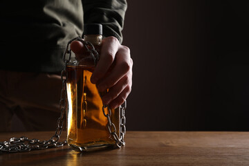 Alcohol addiction. Man chained with bottle of whiskey at wooden table, closeup. Space for text