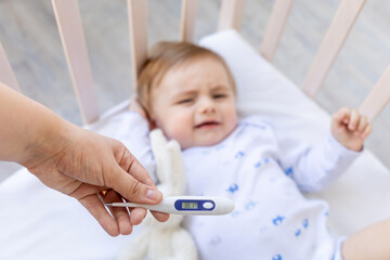 thermometer in focus in mom's hands with a high temperature of 38 degrees on the background of a sick crying baby boy in a crib at home on white bedding, medicine and pediatrics
