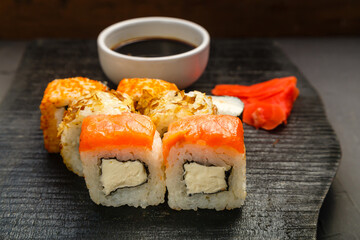 Assorted salmon and shrimp rolls on a black board next to soy sauce and ginger on a dark background.