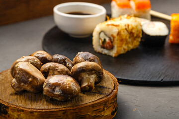 Assorted rolls on a round board next to sticks, soy sauce and baked champignons on a dark background.