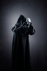 Creepy figure in hooded cloak with human skull mask in hands over dark misty background