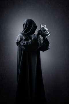 Creepy figure in hooded cloak with wolf mask in hands over dark misty background