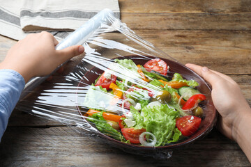 Woman putting plastic food wrap over bowl of fresh salad at wooden table, closeup