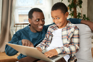 Sly-looking dark-skinned boy kid holding opened laptop on knees, to play games, browse web-sites, smiling sitting next to his eldest brother of 20s, looking at him with smile while pointing at screen - obrazy, fototapety, plakaty