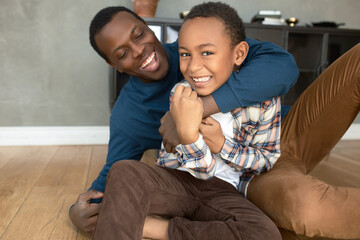 Funny picture of caring and loving dark-skinned dad hugging his misbehaving, naughty boy, both...