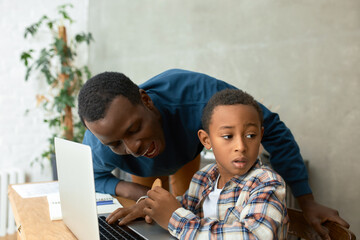 Funny picture of puzzled boy looking aside while his elder 20-years-old brother trying to bite his cookie he's holding in hand, African boy siting in front of opened laptop, studying