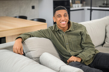 Portrait of carefree young latin guy sitting on the sofa and looking at the camera. Serene multiracial man in casual clothes in relaxed atmosphere spending leisure at home