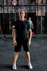 Fototapeta na wymiar Full-length portrait of handsome bald guy in black sportswear posing at camera in gym, looking confident. Caucasian man before workout, cross fit training. sport and fitness concept