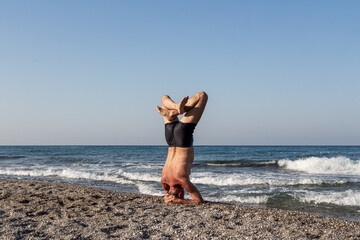Middle age Man in shorts is doing his morning exercises on the beach by the sea. Headstand