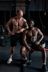 Fototapeta na wymiar Break after hard cross fit training. Portrait of two young sporty men having rest in gym after workout. Diverse interracial mixed-race male fitness models posing at camera, having strong muscular body