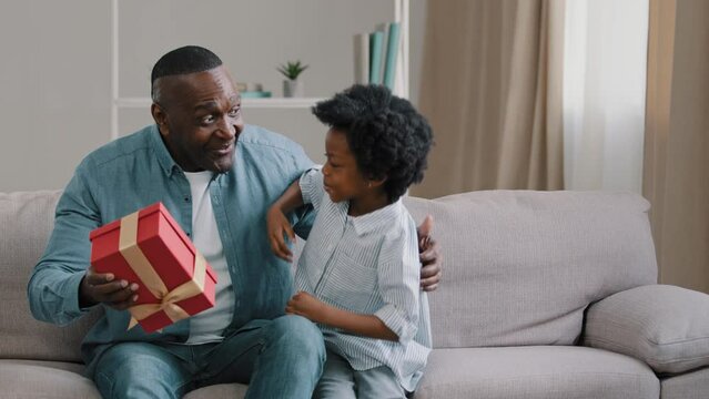 Little african american loving daughter hides box gives gift to beloved daddy on birthday or father day congratulates parent surprised happy dad sitting on sofa hugging kid girl strong family embrace