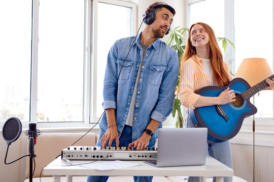 Nice caucasian Male music teacher giving instructions to student female learning to play guitar during home music lessons, performing music together,using piano and laptop, smiling, enjoying