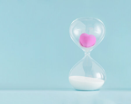 2022. Minimal abstract creative concept with pink heart stuck in hourglass on isolated pastel blue background. Eternal or imperishable love idea. Gift or greeting card for wedding or Valentine’s Day.