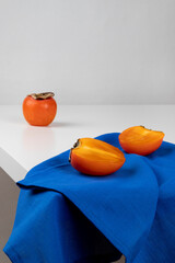 A cut ripe persimmon is lying on a blue towel. White table, close-up, vertical orientation and copy space.