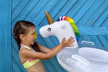 A girl in a swimsuit and yellow sunglasses holds a unicorn-shaped swimming circle with a rainbow mane in front of her. A child with a tan from the hot summer sun is getting ready for swimming