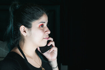 Obraz premium beautiful latina woman with green hair, receiving a call on her cell phone, with a stressed expression. girl sitting in her room talking to her friends. very dark black background.