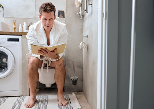 Male reading book while sitting on toilet bowl at home, relaxing, having rest. Young guy in domestic bathrobe in the morning, in broght cozy bathroom. lifestyle, people, everyday life