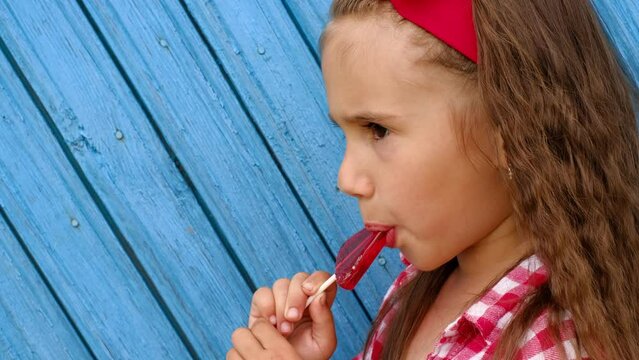 The child eats and licks a lollipop, enjoying the taste of candy. Profile of a girl with a sweet in her hands on a blue background of a wooden wall made of planks