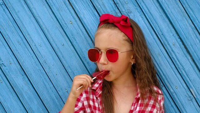 Girl in red sunglasses holds a triangular lollipop on a stick in hands, licking it with tongue. Profile of a child who eats candy on the background of a blue board