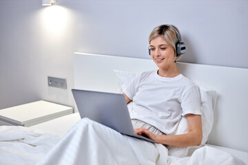 happy caucasian woman posing enjoy music at headphones, using laptop sitting on bed write messages,listen to music. lady in domestic outfit relaxing, having rest at home alone, leisure